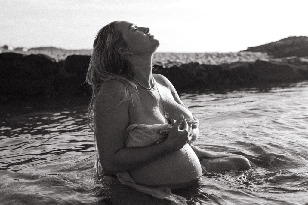 A pregnant mother sits in the water on a beach , her last trimester belly is outlined in the sunrise. She is looking up while the water surrounds her pregnant belly