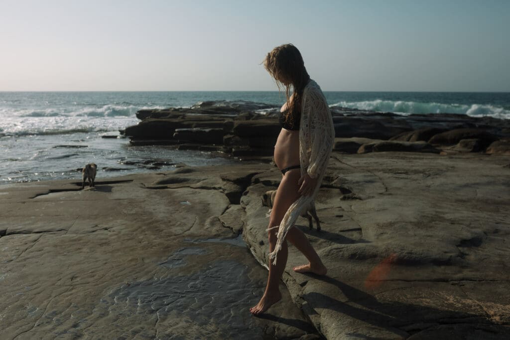a mother holds her pregnany belly while walking on a sunrise beach , she is stepping on rocks and the sunrise lights up her whole body
