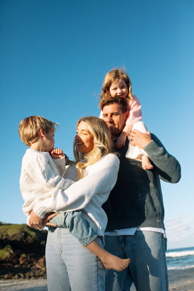 Mother, father and children on cabarita beach , the little girl is balancing on her dad shoulders and boy balanced on his mothers hip. They are smiling while the setting sun is lighting up their cheerful faces 
