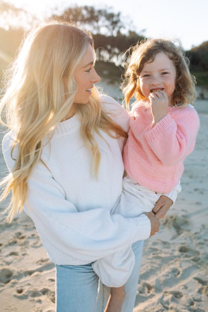 Mother and child on Cabarita beach , they are being lit by the setting sun . The daughter is grinning at the camera while mother cuddles her