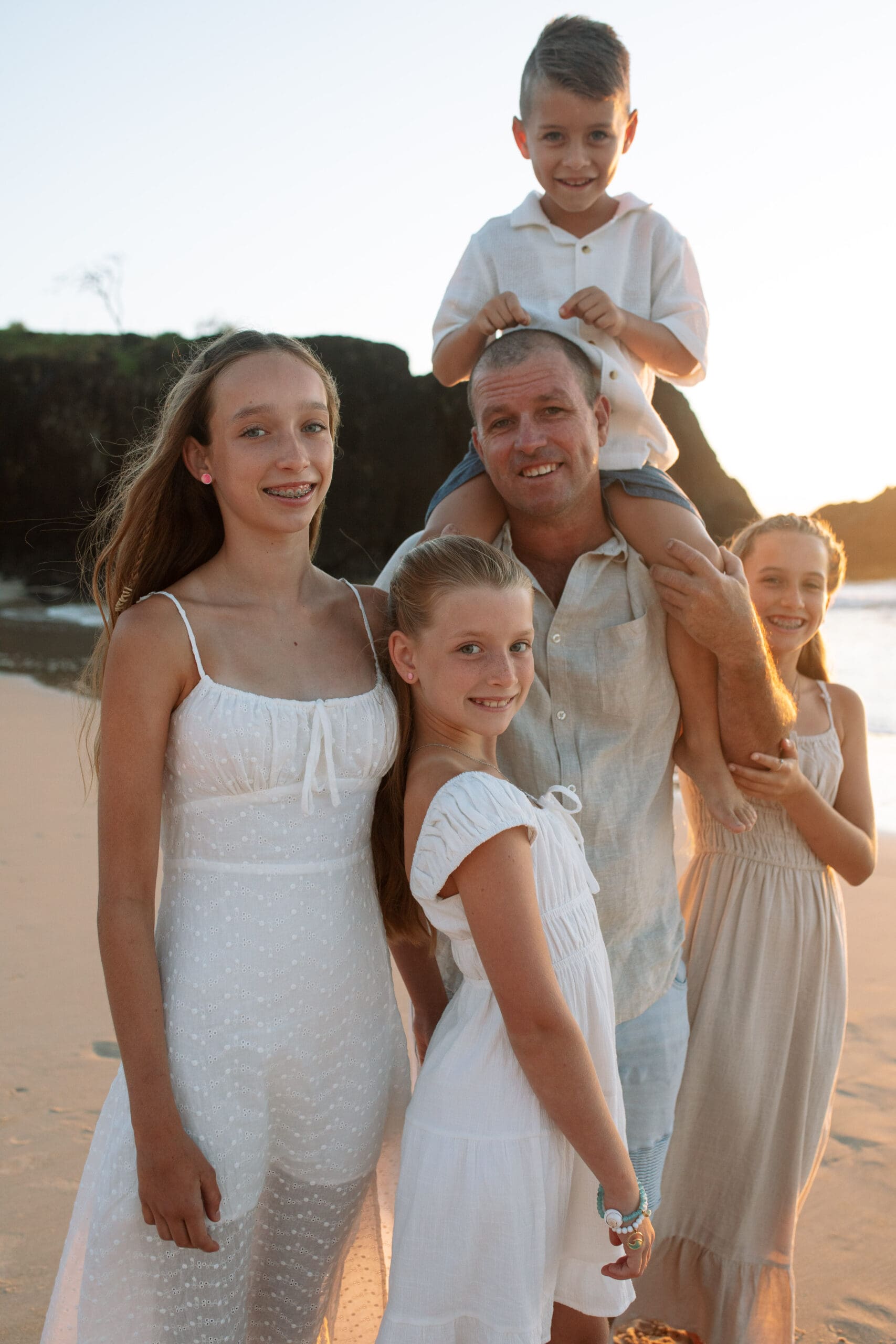 father and children all joyfully smiling , you can can see Fingal Headland in the backround. They are on Dreamtime BEach in the sunruse light , all weraing neutral colours