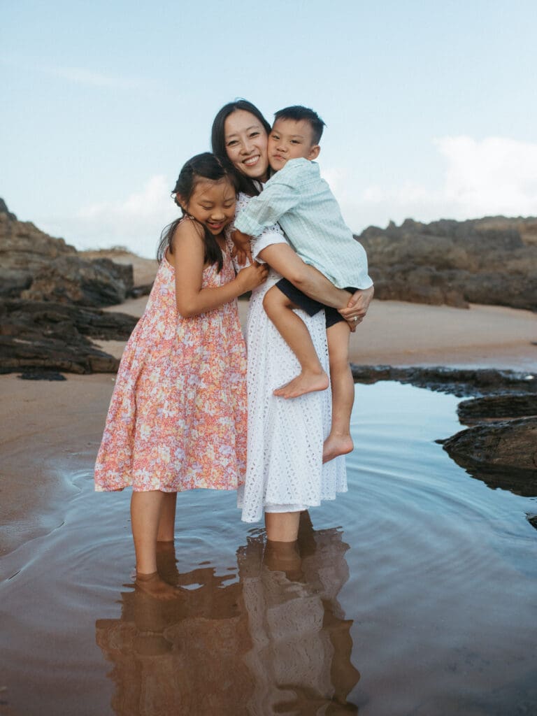A mother is holding her son and being squeezed by her daughter , they are joyfully looking into the camera and standing in a reflective pool of of water in a natural beach family photo on the gold coast
