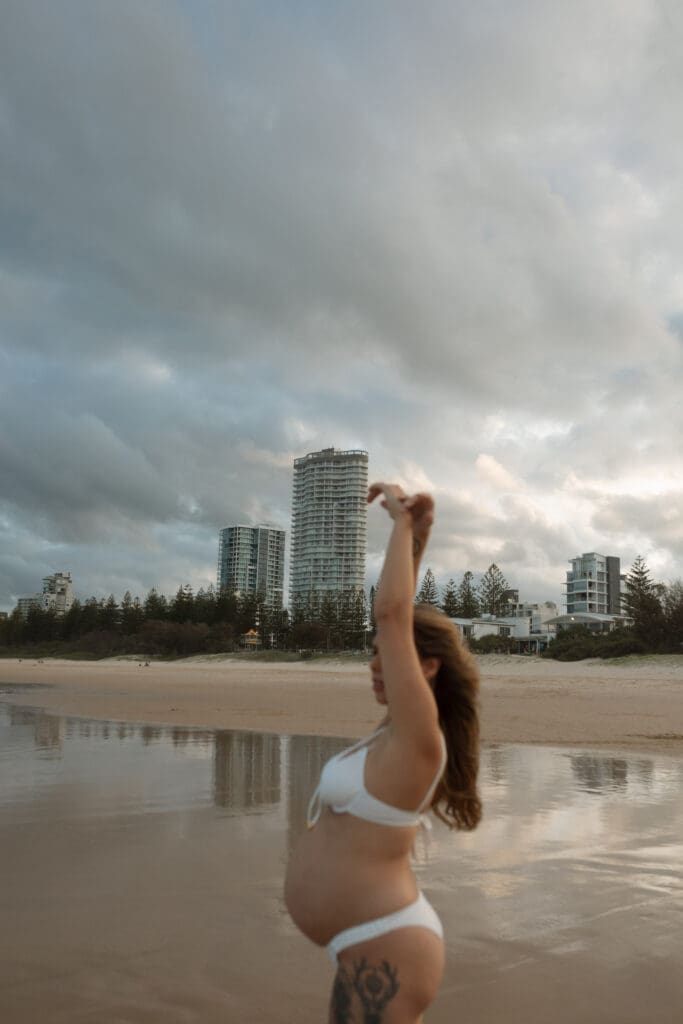 An expectant mother reaches to the dramatic skies against the Burleigh cityscape . Her belly is bare and she is on the beach