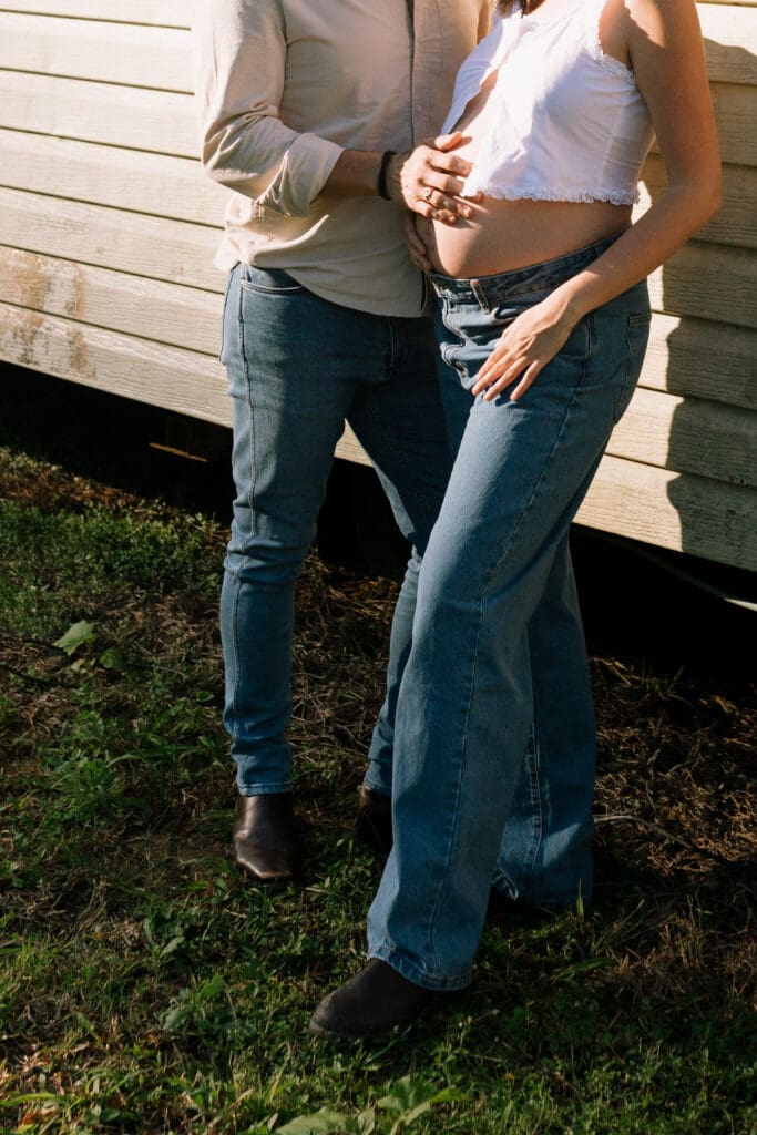 A couple cradle a pregnant belly in direct golden sunlight , they are standing against a building on a farm in the Hinterland
