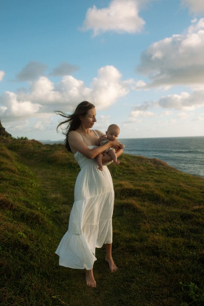A mother holds her baby at Lennox Heads , the wind is blowing her hair and dress . There is golden sunrise and a natural and candid family photo on the Headland
 
