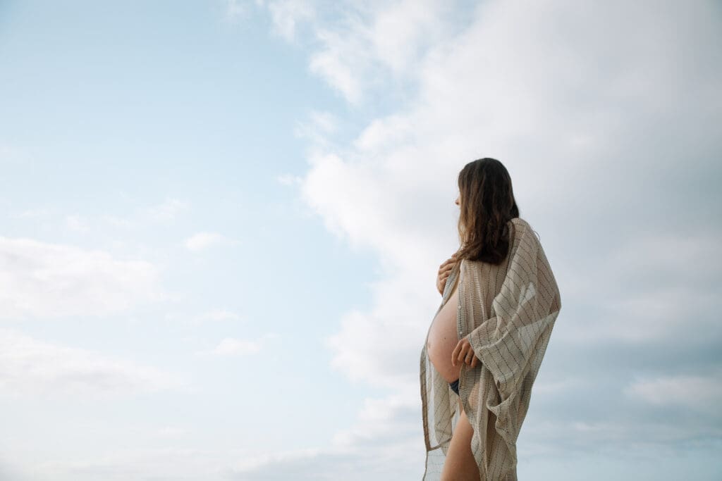 A pregnant mother stands against the sky and clouds  caressing her belly on a Tweed Heads Beach in the Northern Rivers 