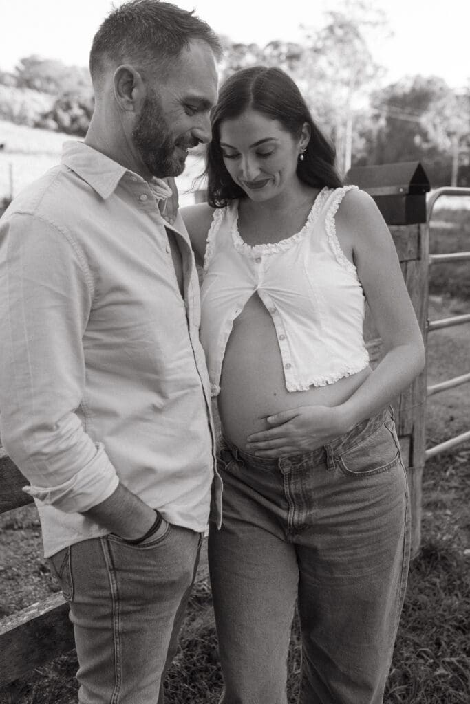 A mother and father cradle a pregnant belly , they stand against a fence in the coastal hinterland im the afternoon sun
