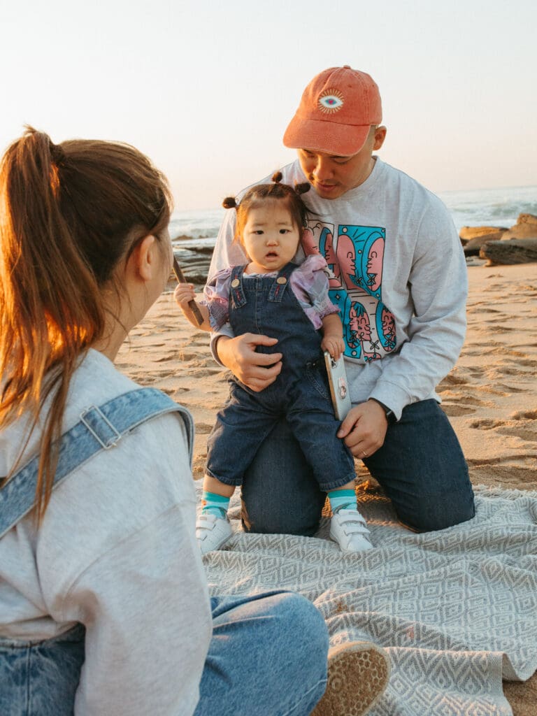 Mother , father and toddler playing on a Gold Coast Beach at sunrise. The toddler is looking at the camera and framed by her mother and being by her father. It is a light filled image.

