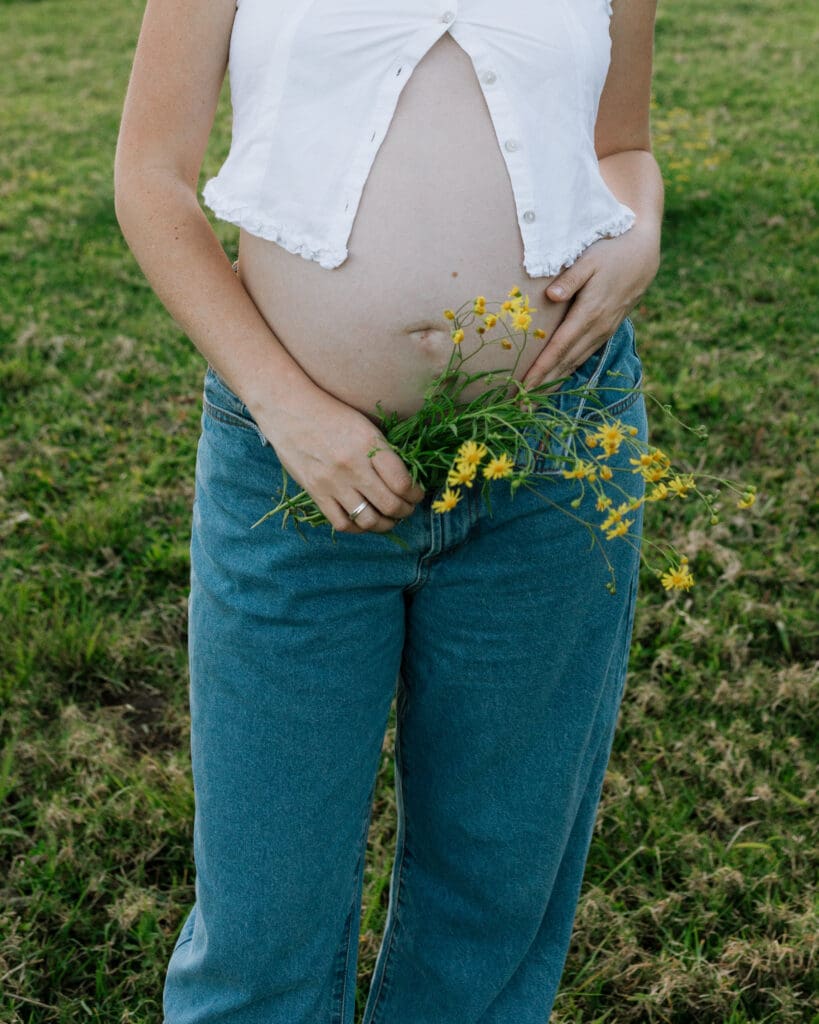 A mother holds some yellow flowers against her belly , she is standing in a coastal hinterland field in natural subtle light