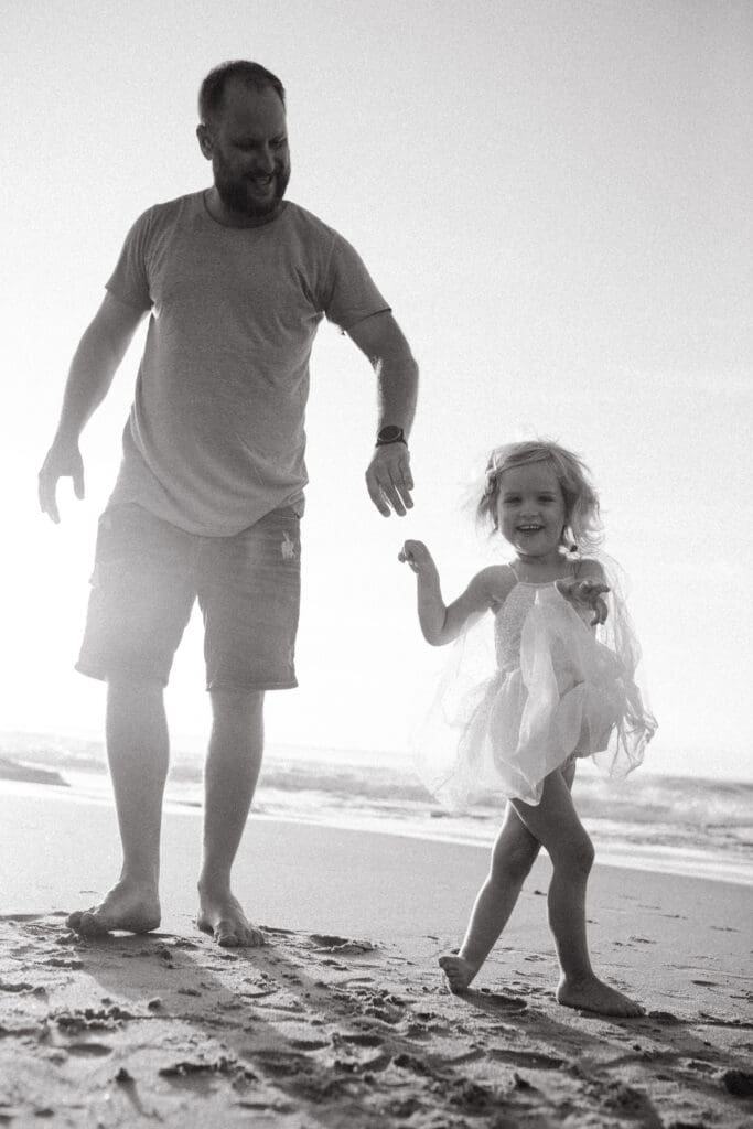 A dad and his daughter happily play on the beach , the daughter has a tutu on and is twirling and giggling. The ocean is in the background , the waves lit up by the rising sun. 