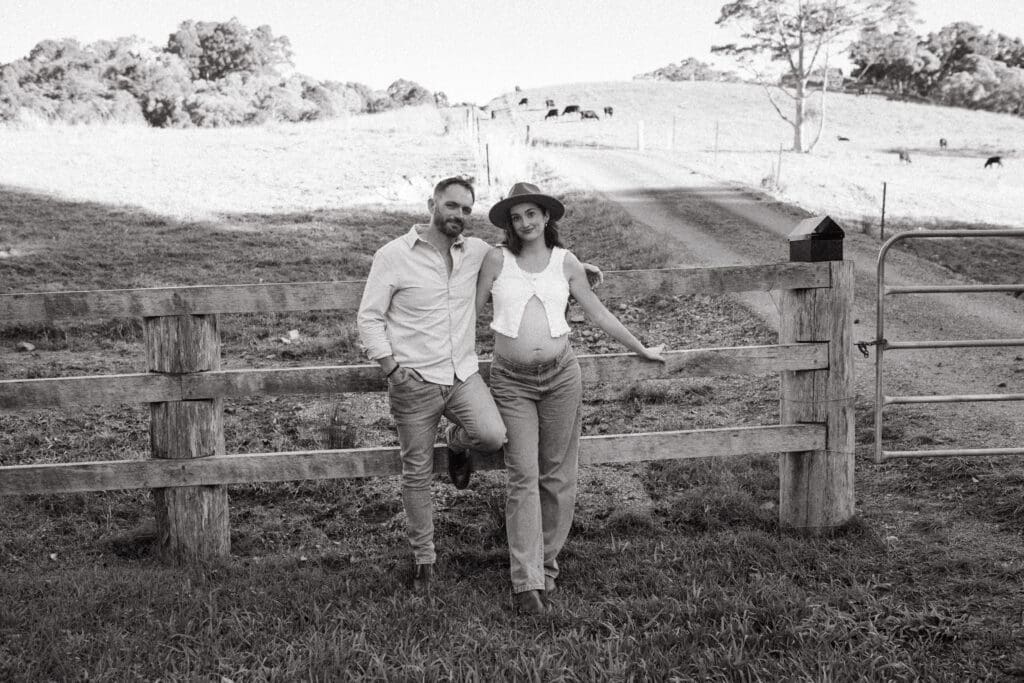 A couple lean against a fence in the Hinterland, cows in a field in the background and the mothers pregnant belly is defined