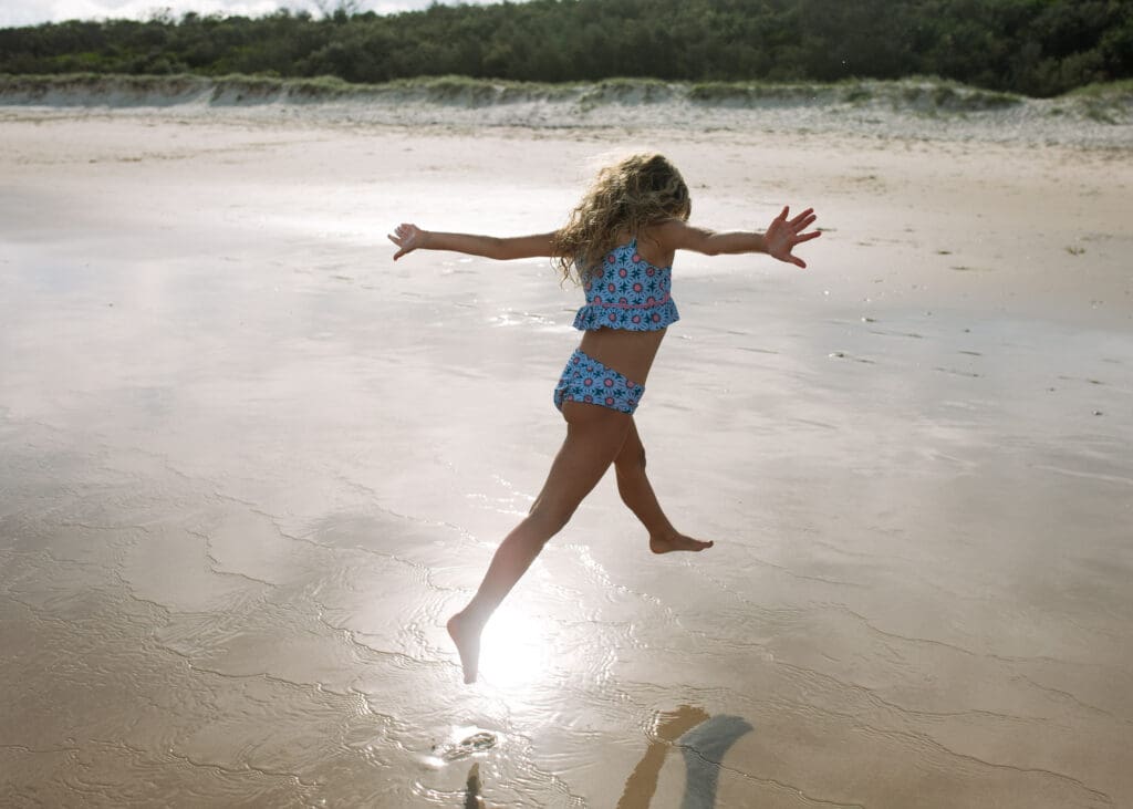A girl leaps and jumps in the setting sun at Hastings Point, the suns reflects off of her and and is golden. The sand dunes can be seen in the background shadows. 