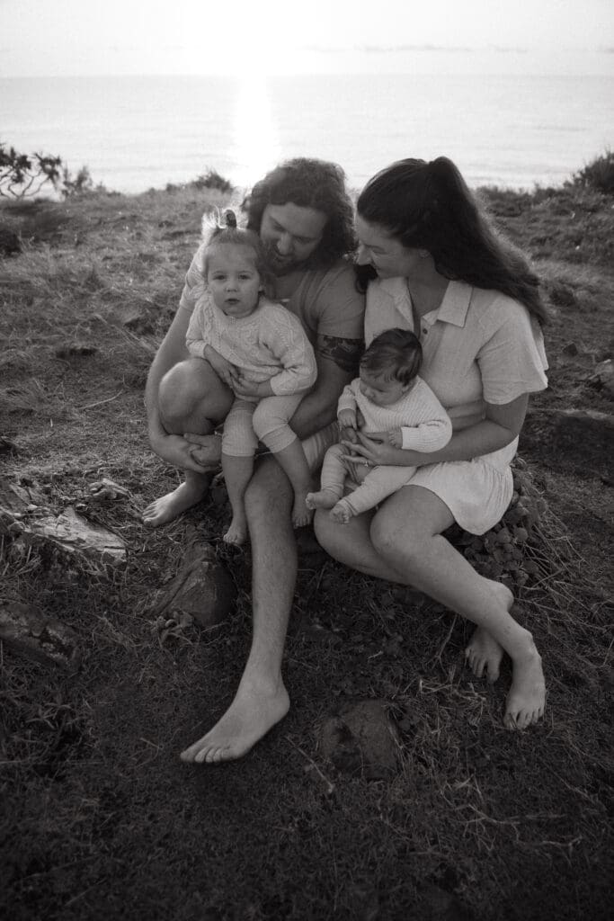 Family of four cuddling on the cabarita headland, they sit close togetherwhispering while the toddler grins at the camera