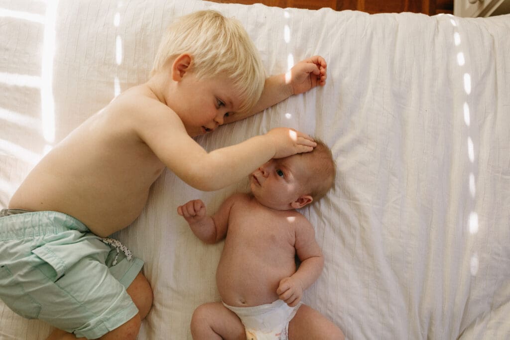 A newborn baby and toddler sibling play on a bed in a bright kingscliff home