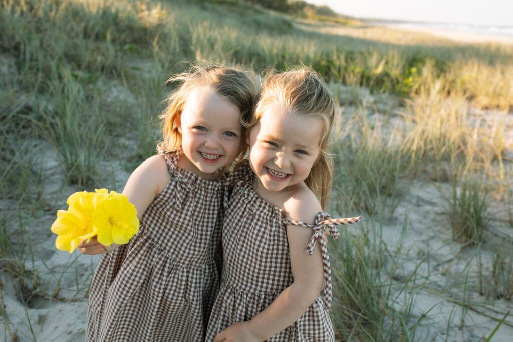 Two toddler twins smile cheekily at the camera , they are holding a bright yellow flower and are cuddled close in the sand dunes at golden hour at Cabarita Beach