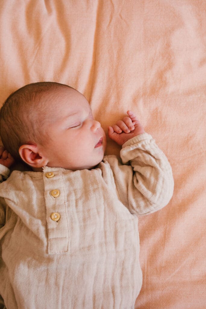 A sleeping newborn in a gold coast home , tiny hands are curled up and  eyes are closed . The baby sleeps on soft wrinkled linen