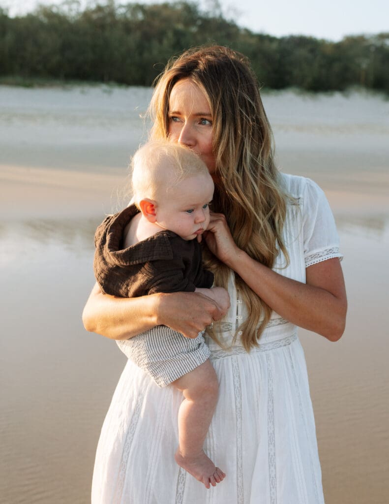A mother holds her baby tightly on the beach at Fingal Head , she is outlined by golden hour light kissing her sons head