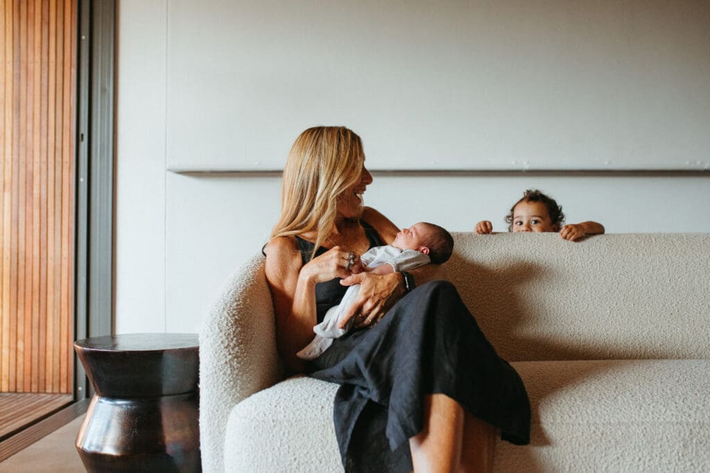 A grandmother holds her newborn baby in a Gold Coast home , her toddler grandchild peeps out from behind the couch , she is smiling at the toddler
 while cuddling her new grandchild