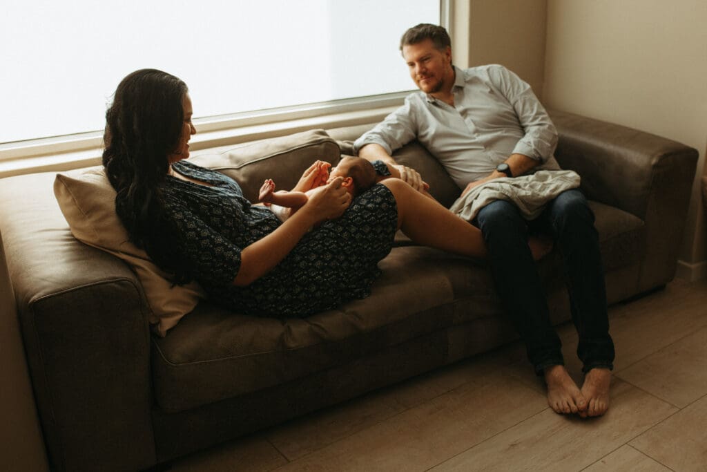 A mother rand father sit by a window in a gold coast house , the light from the window lights up the awake newborn baby on the mothers lap