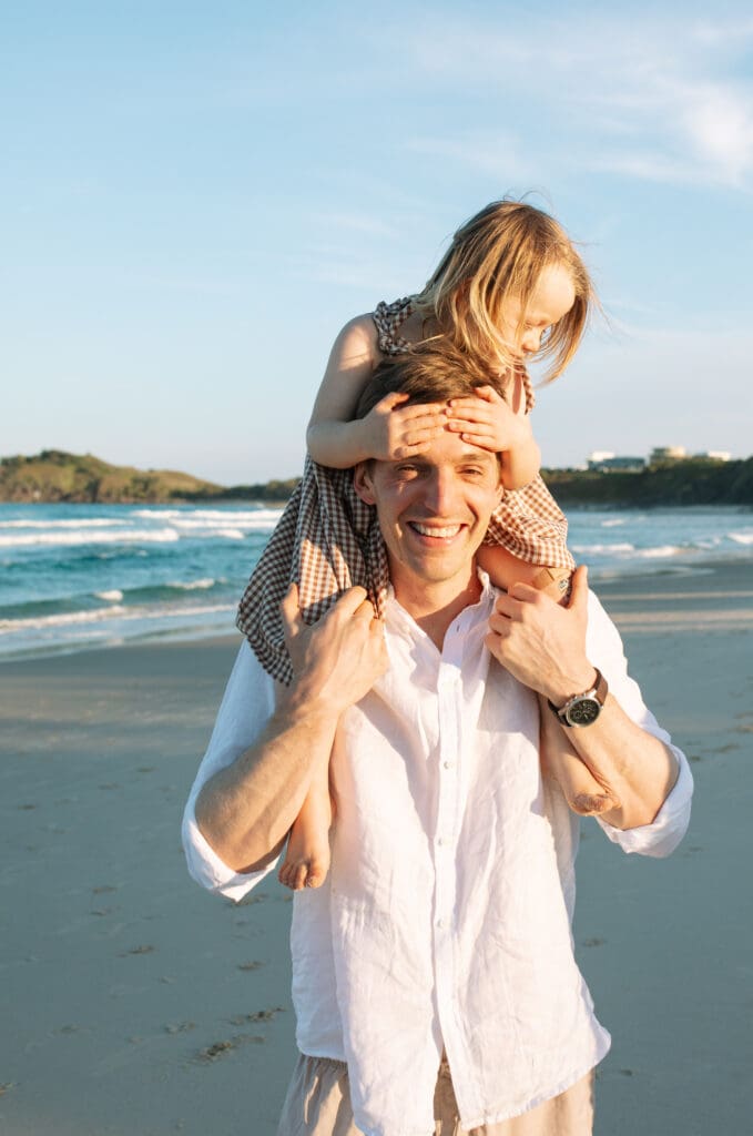 A toddler girl balances on her dads shoulders , he is looking at the camera and smiling. Cabarita ( Norries Headland) can be seen in the background and 
lit up by golden hour light