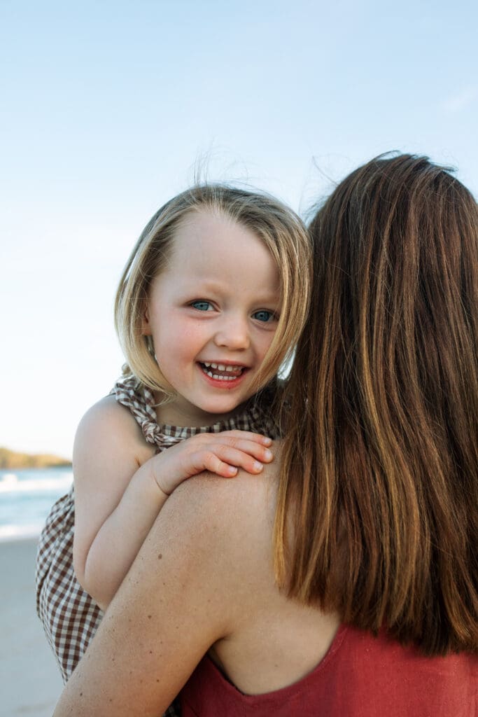 Toddler on her  mothers back smiles joyfully at the camera at cabarita main beach at golden hour