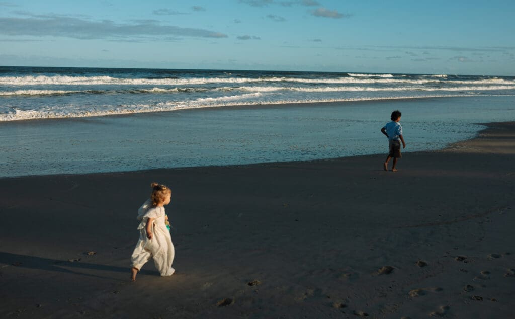 A toddler and her brother race in the sunlight on cabarita beach at sunset , the waves are golden and shadows are soft , the toddler is in the light and her brother in shadow