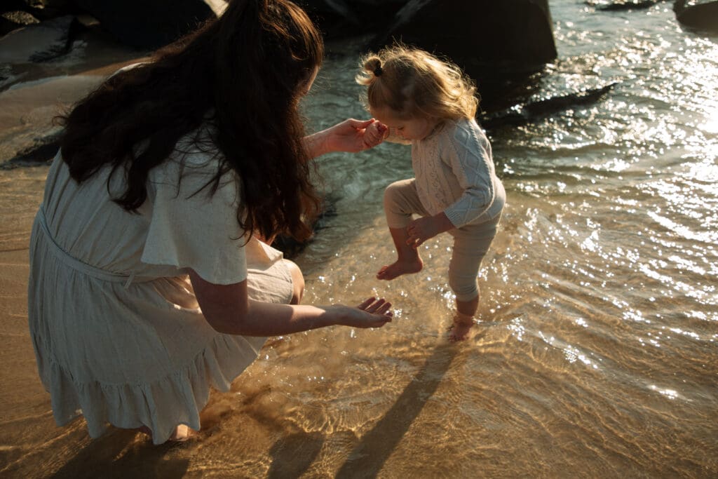 A mother plays with her toddler in the morning sunrise at Cabarita Beach . The toddlers giggles and kicks cold ocean water and the sunlight glitens in the sunrise