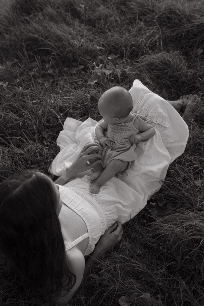 A mother lies in the grassland on the lennox heads headland and cuddles her tiny baby. she plays with her baby's hands and toes and sunrise lightly illuminates them
