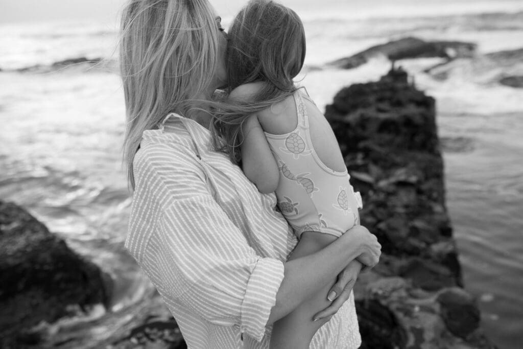 A Mother hugs her daughter tightly on the beach , she is kissing her daughters head with her eyes closed. They are standing on a quiet northern rivers beach and the wind is gently lifting their hair