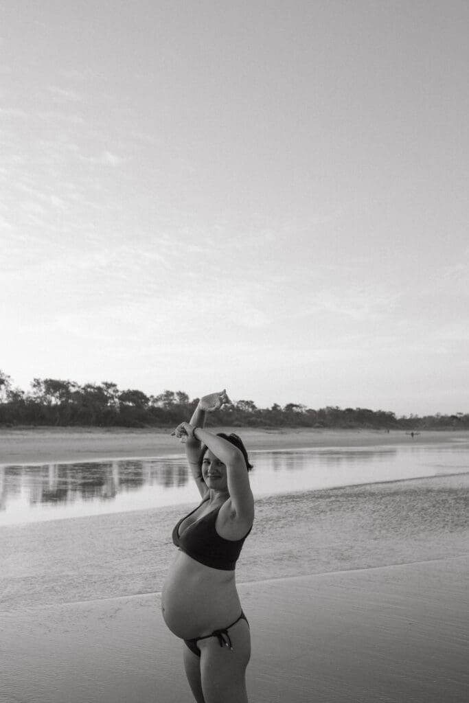 A heavily pregnant mother lifts her hands to the setting sun at Broken Head Beach , the trees in the beach dunes in the background are reflected in the clear water