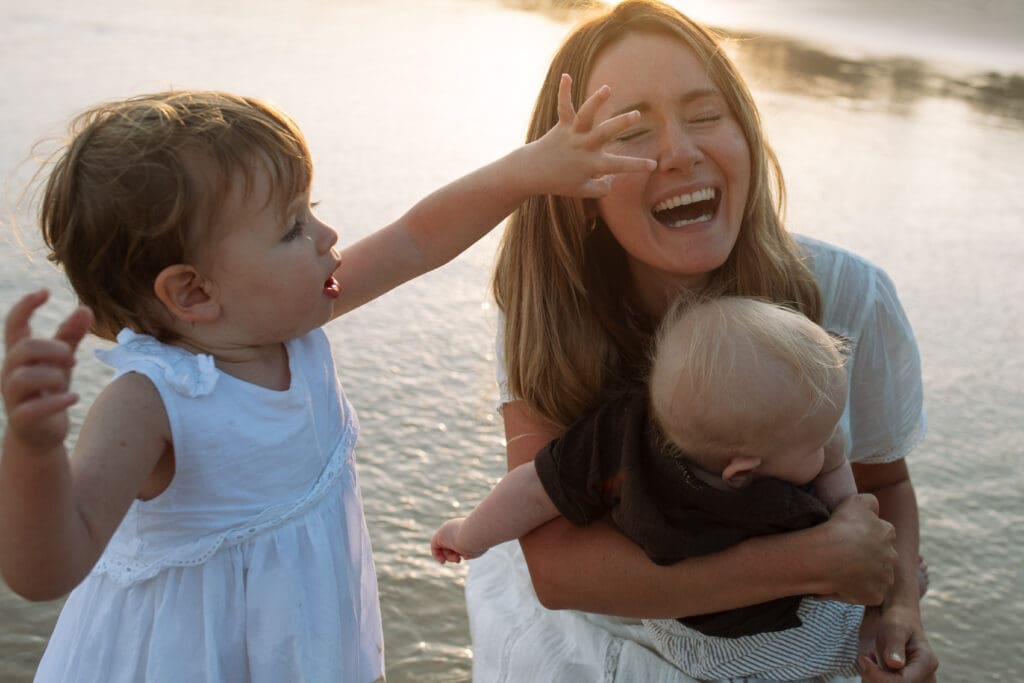 toddler toddler touches her mothers face. Her mother is giggling , the beach at dreamtime can be seen glowing in the background
