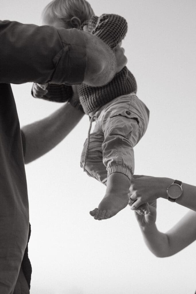 A father lifts his toddler in the air and mother dusts beach sand off his little toddler toes in a natural moment