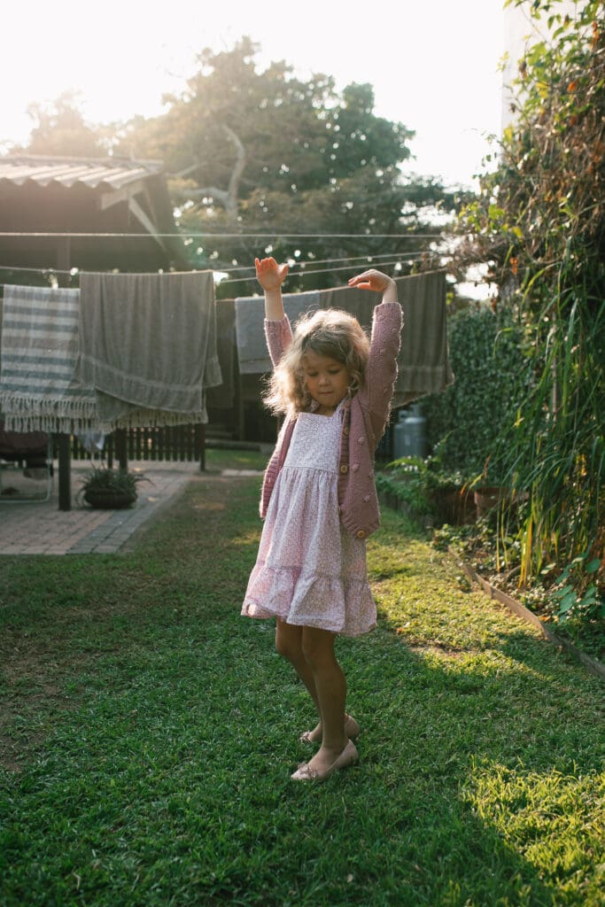 A girl stands in her backyard and is lifting her hands up in gentle morning light , she is natural 