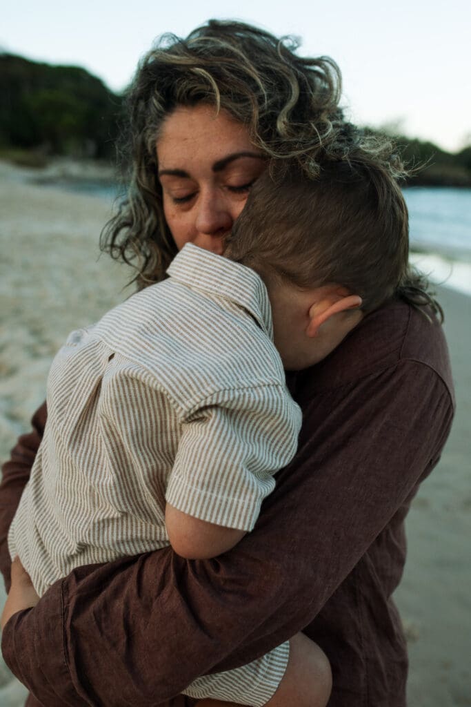 A mother holds her toddler tightly and is softly lit by the setting beach sun , the Pottsville creek  beach is in the background