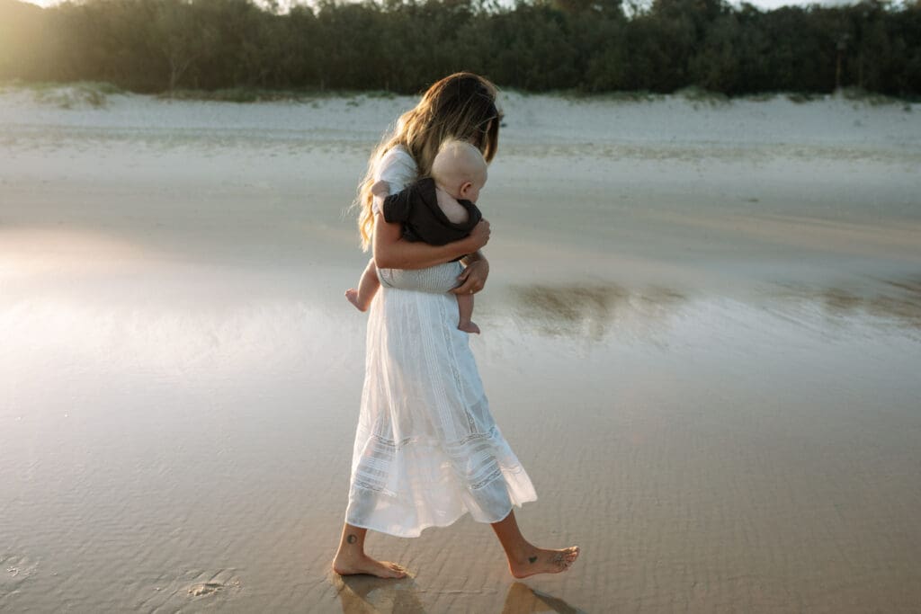 A mother holds her baby in the sunset light at Dreamtime Beach at Fingal in the Northern Rivers , the golden light is refelcting off the sand and golden dunes in the background 
