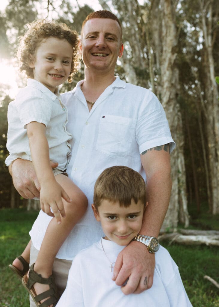 A dad hold his boys while laughing at the camera , the boys are staring  into the camera , the setting sun lights up their hair and gives a  sun flare that feels nostalgic . They are standing in a paddock on Round Mountain Road

