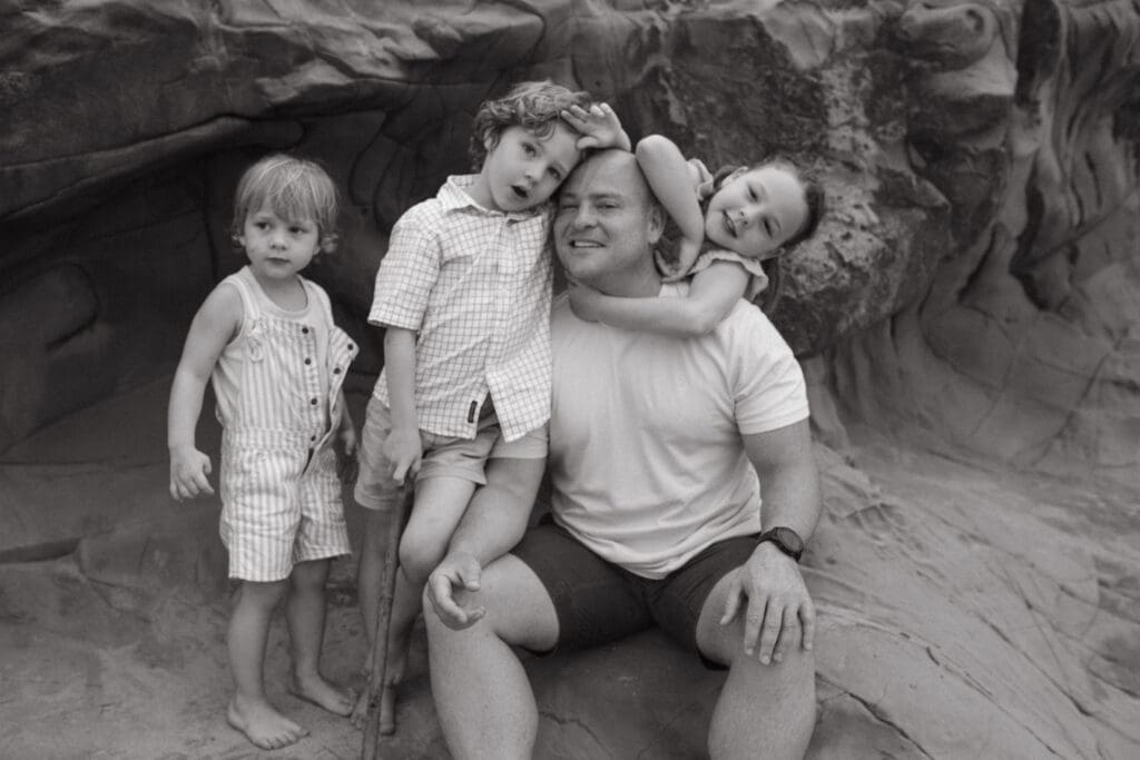 Children climb over their dad while he stares at the camera with a cheeky grin
