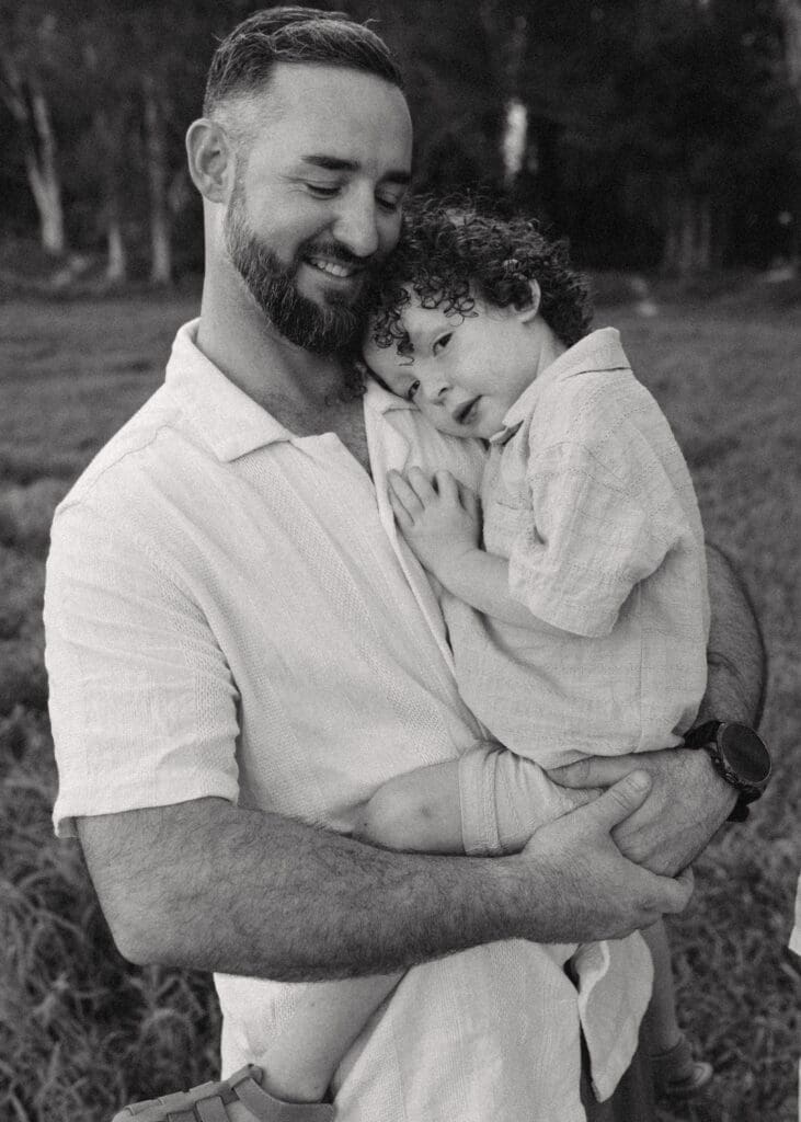 A father and sun cuddle in a hinterland paddock, the child clutches at his dad whose eyes are closed, the toddler is staring at the camera  .Bush and Blue gums are in the background