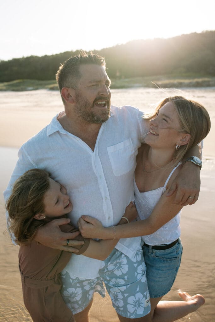 A dad and his two daugers play in the waves at Kingscliff beach , they are lit up by the setting sun , it is a candid moment with him squeezing his daughters and them lovingly giggling
