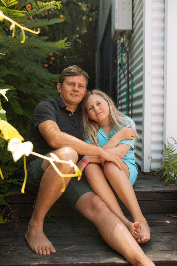 A dad and daughter sit on a backyard step in a Lennox Heads Air BnB, they are staring at the camera framed by a vine , barefoot and casual