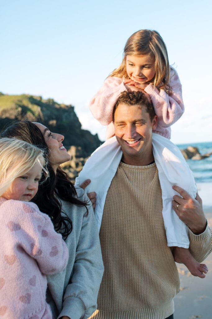 A dad holds his young daughter on his shoulders while his wife gazes up. Norries headland is in the background
