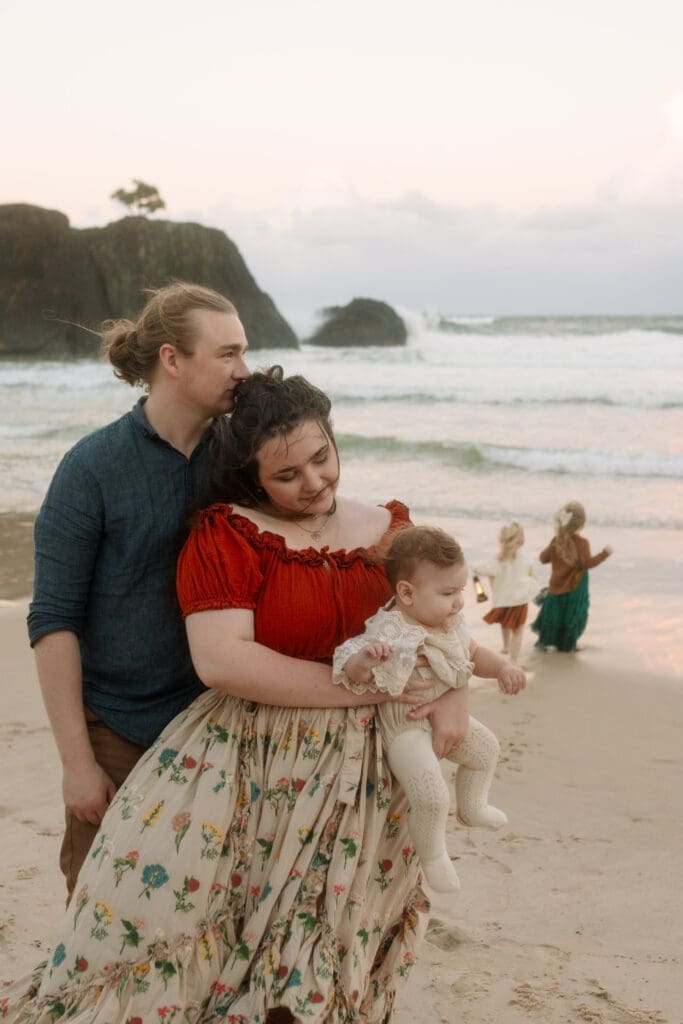 A father kisses his wives forehead while their toddlers play in the waves at Dreamtime Beach. The wife is holding and looking at the baby