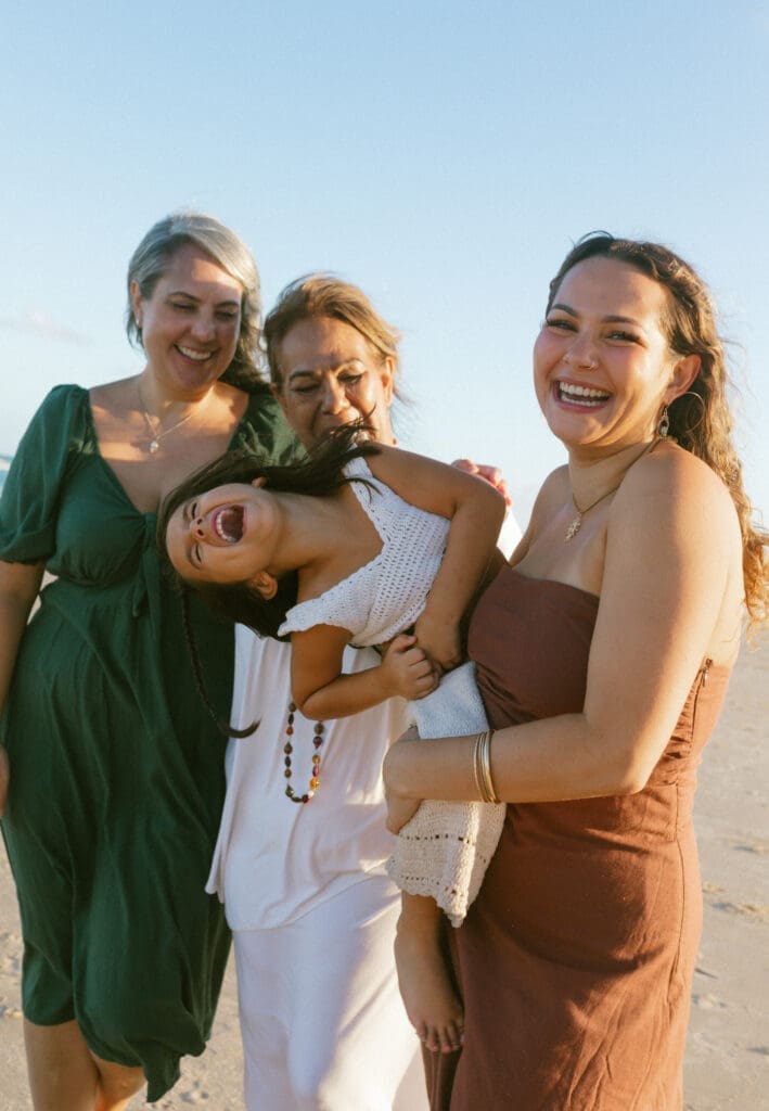 a little girl twists and giggles wildly in her aunts arms while her granny look son . They are standing on a kingscliff beach
