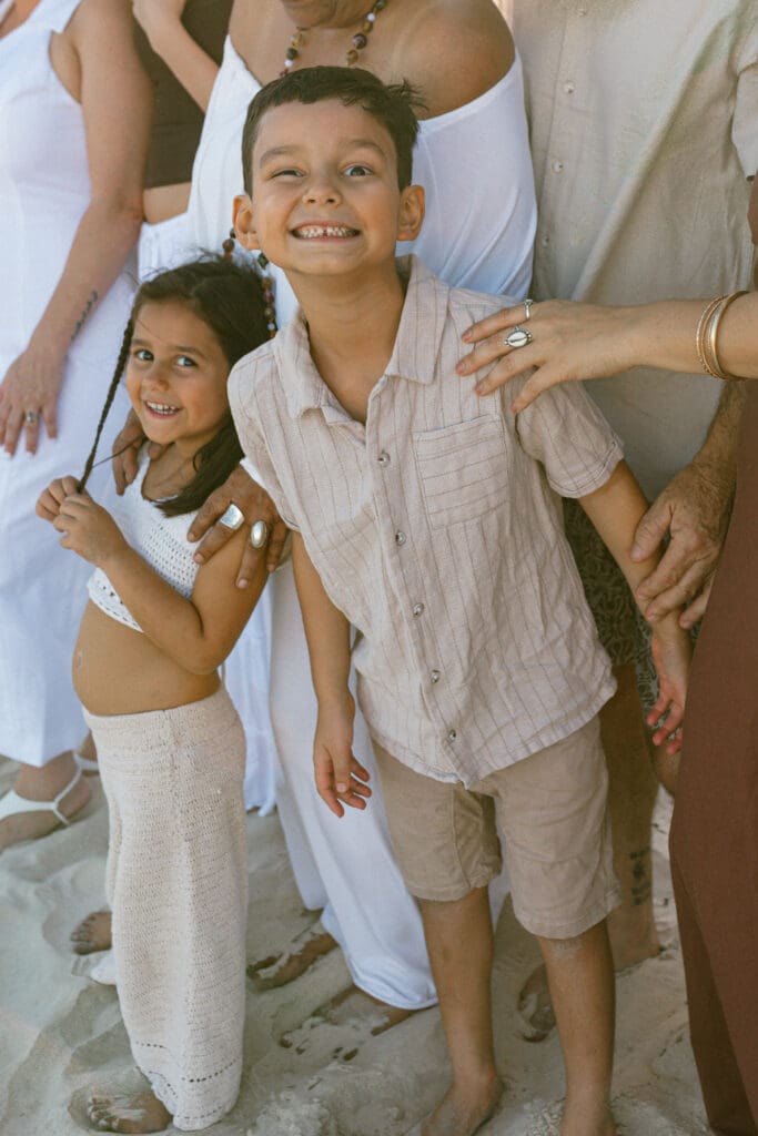Two young siblings laughing together with grandparents' hands on their shoulders during a candid family beach photo in Kingsclif