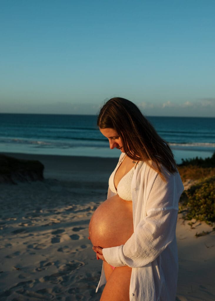 Pregnant woman holding her belly beside the ocean during a maternity photoshoot at Belongil Beach