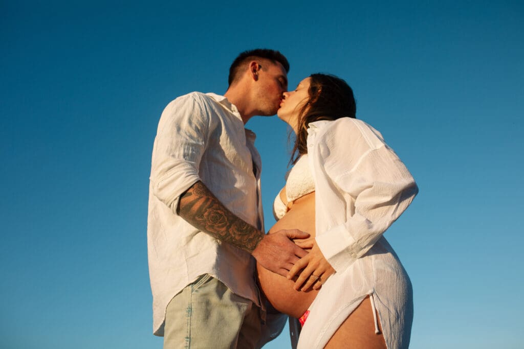 Expectant couple kissing with their hands on the baby bump during a maternity photoshoot at Belongil Beach, Byron Bay
