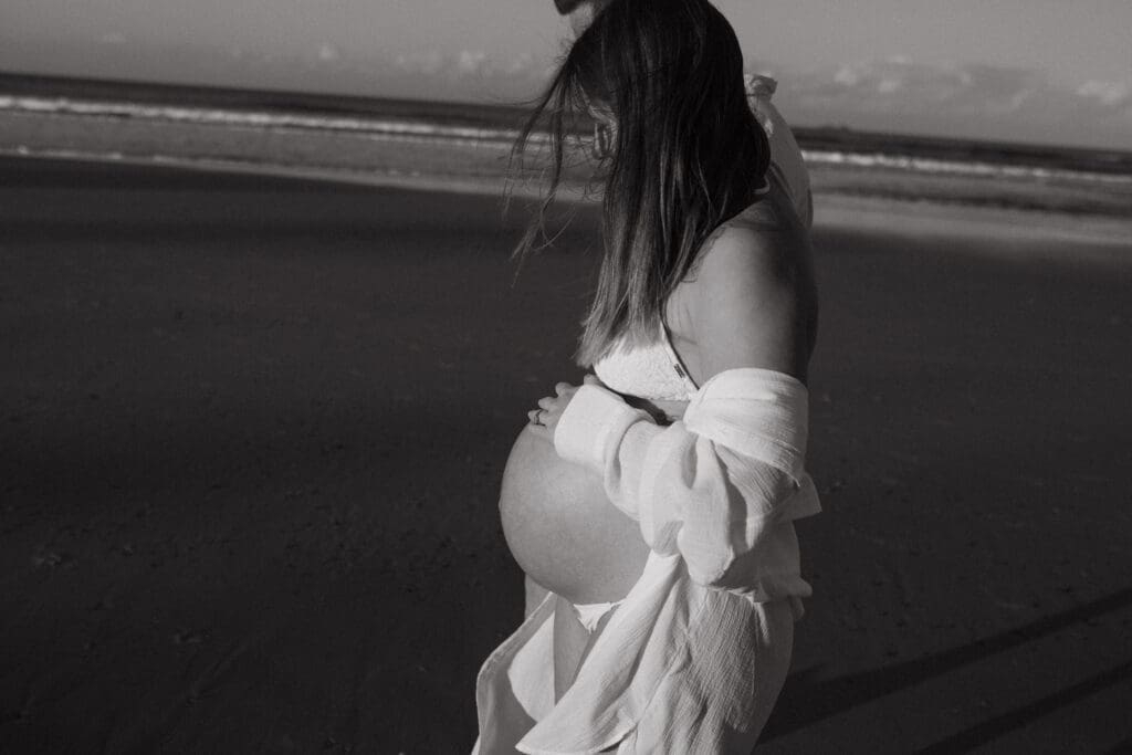 Candid black-and-white portrait of a pregnant woman walking along Belongil Beach during her Byron Bay babymoon