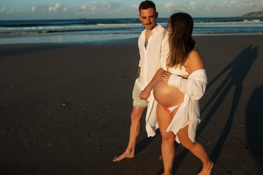 Expectant couple walking barefoot together during their Belongil Beach maternity session in Byron Bay.