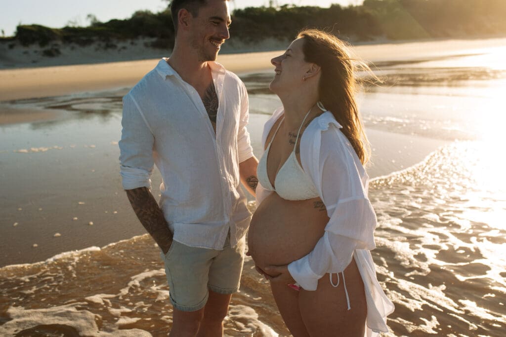 Expectant couple laughing together beside the water during their relaxed Byron Bay babymoon photoshoot